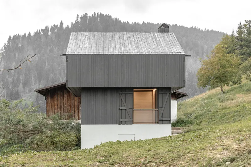 Dark timber-clad volume with gabled roof and folding shutters on white base in alpine meadow