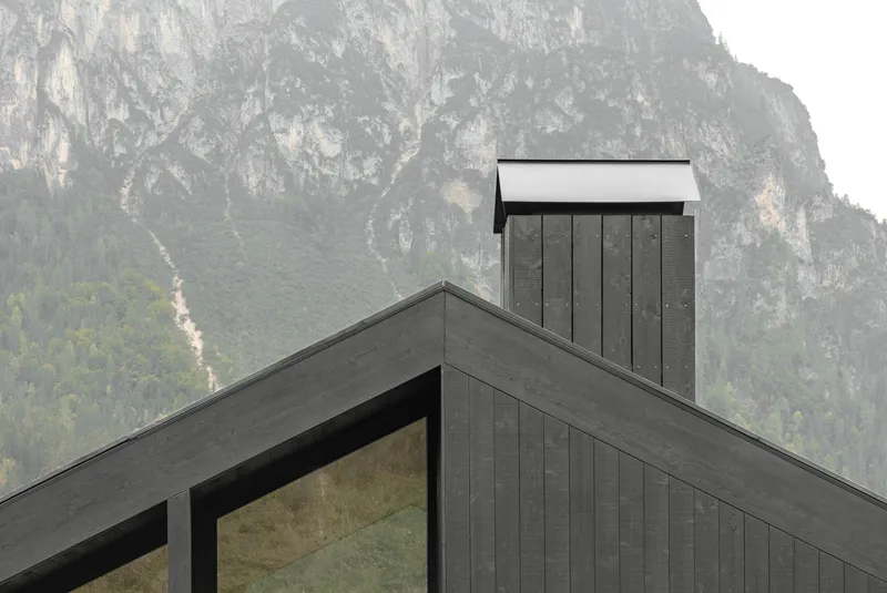 Vertical chimney clad in dark timber rising above the gabled roofline against a rocky mountain face