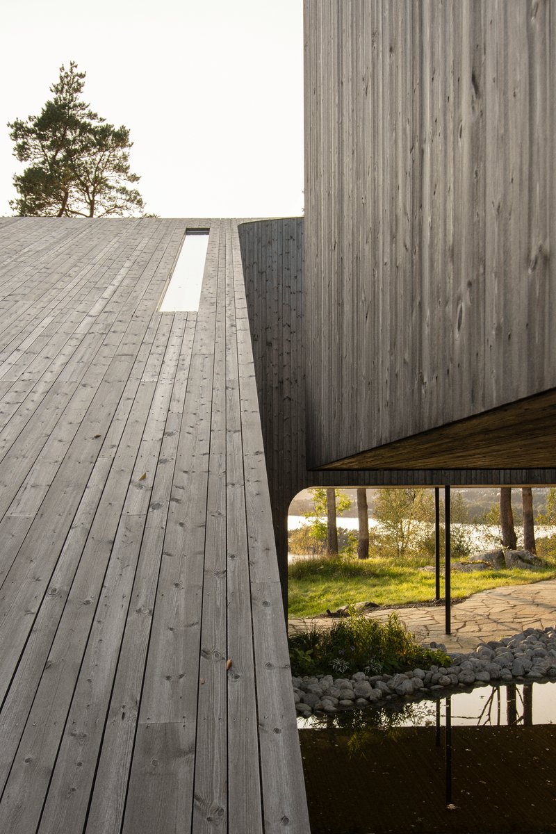 Timber deck wrapping around the vertical-clad volume above a columned terrace and pond edge