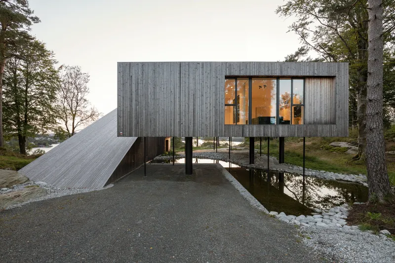 Entry view of the elevated timber box and sloped roof with illuminated interior at dusk