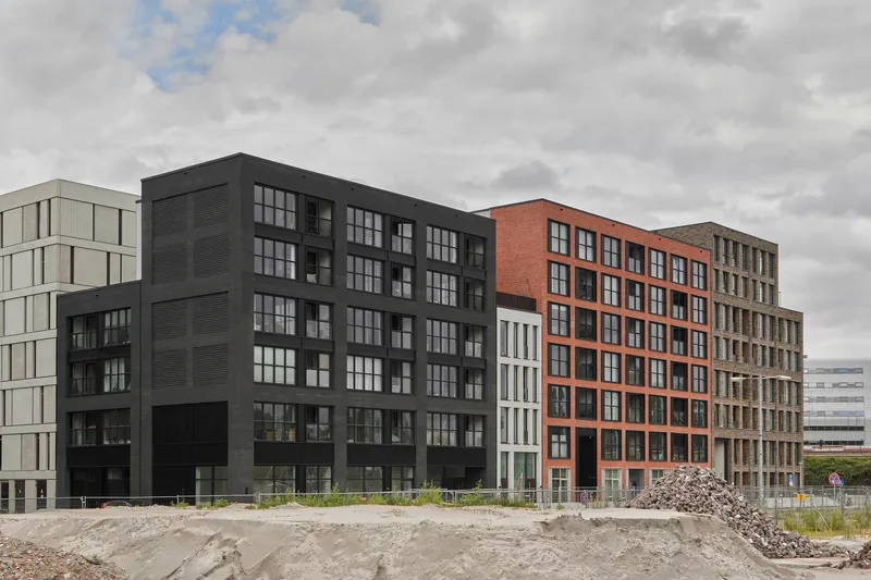 Street view of three multiunit blocks with orange brick, black cladding and grey panel facades across undeveloped ground