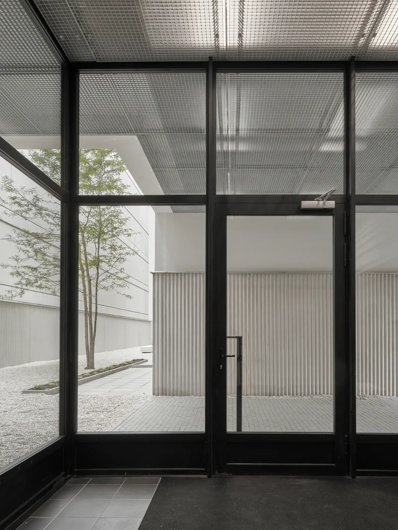 Black-framed glass entry doors with translucent corrugated overhead canopy and gravel courtyard beyond