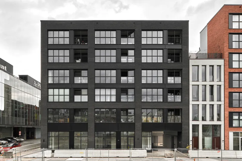 Black cladded facade with large gridded windows and recessed balconies flanked by neighboring buildings