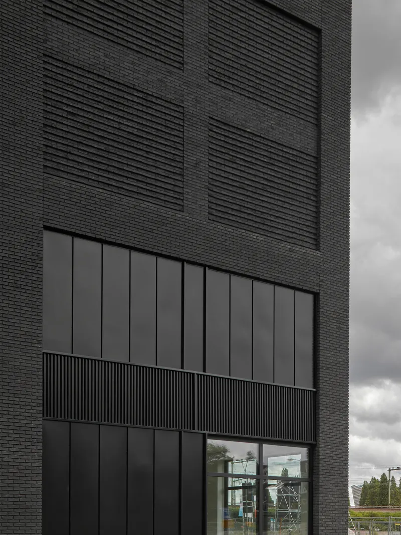 Dark brick facade with recessed balcony and horizontal corrugated metal panel beneath clouded sky