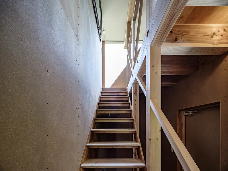 Timber stair ascending toward a bright skylight between concrete wall and cross-braced wooden railing
