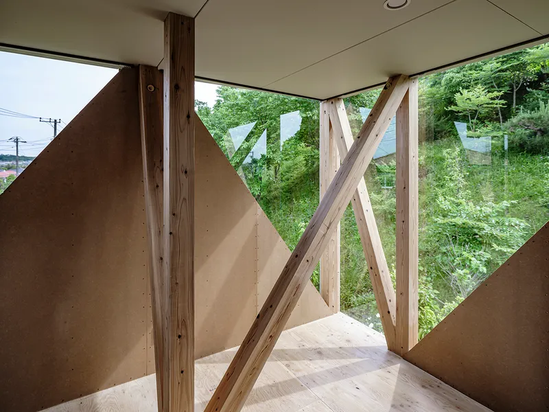 Interior corner with diagonal timber bracing framing a view of dense green foliage outside