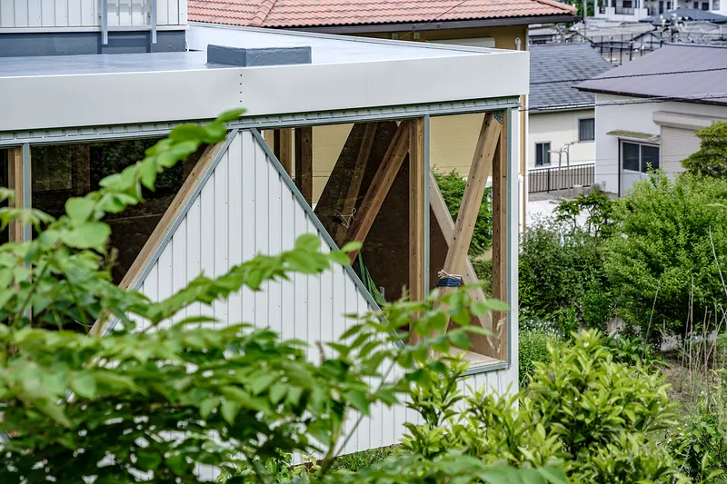 White corrugated metal facade with exposed diagonal timber bracing visible through a glazed opening surrounded by vegetation
