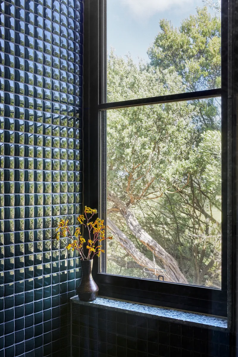 Bathroom window sill with dark mosaic tile wall and yellow flowers in vase overlooking trees