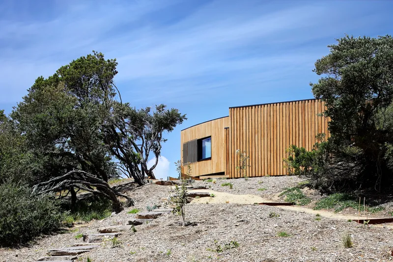 Vertical timber-clad volume perched on a gravelly slope with wind-shaped trees flanking both sides
