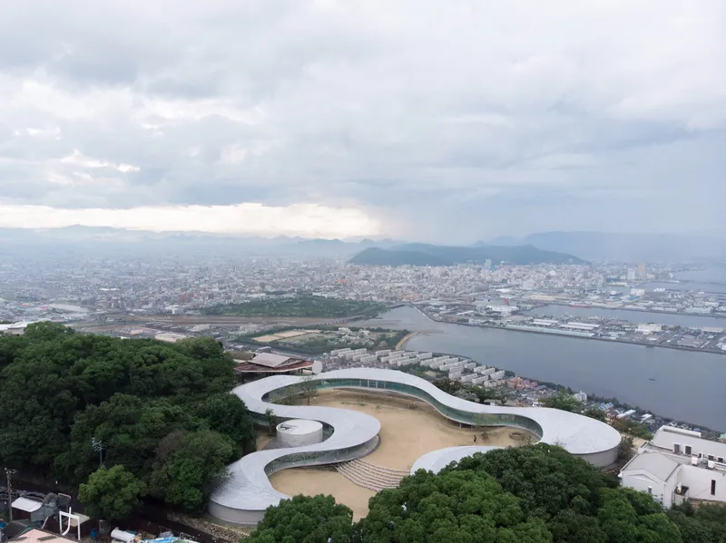 Aerial view of the spiraling white roof nestled among forested hillside overlooking a coastal harbor