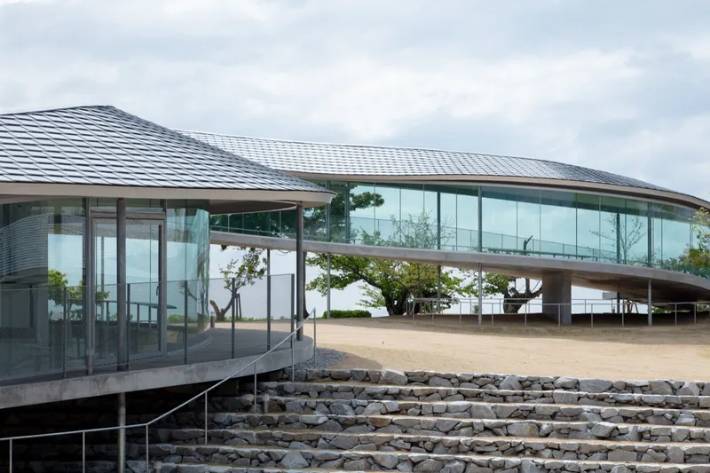 Stone amphitheater steps leading up to the glass-walled pavilion beneath its rippling metal roof