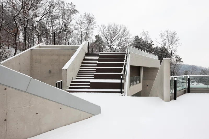 Exterior concrete stair ascending from a snow-covered terrace with bare winter trees in the background