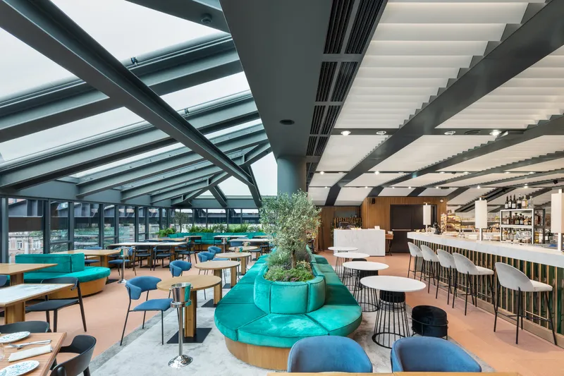 Rooftop cafe area with planted circular seating and folded ceiling baffles opening to glazed perimeter views