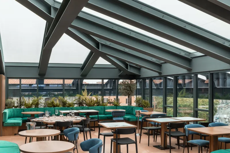 Dining area beneath exposed steel roof trusses with skylights and turquoise banquette seating