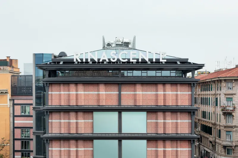 Facade with terracotta panel cladding and horizontal glazing strips beneath a dark rooftop structure under overcast sky