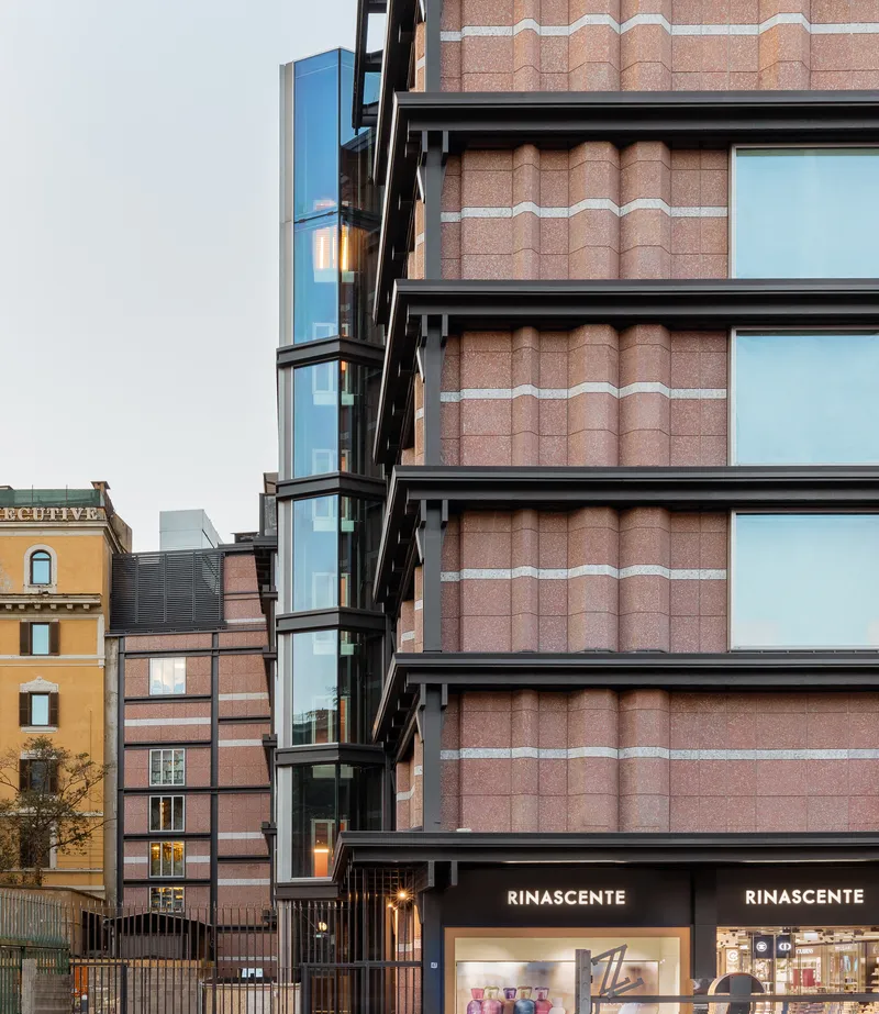 Street view of stacked blue glass balconies beside terracotta paneled facade with horizontal glazing at twilight