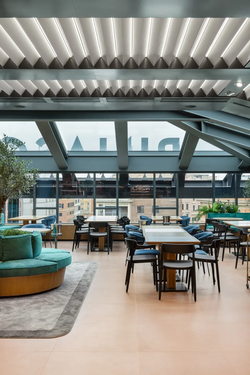 Dining area with folded ceiling baffles and steel trusses framing glazed walls overlooking neighboring rooftops