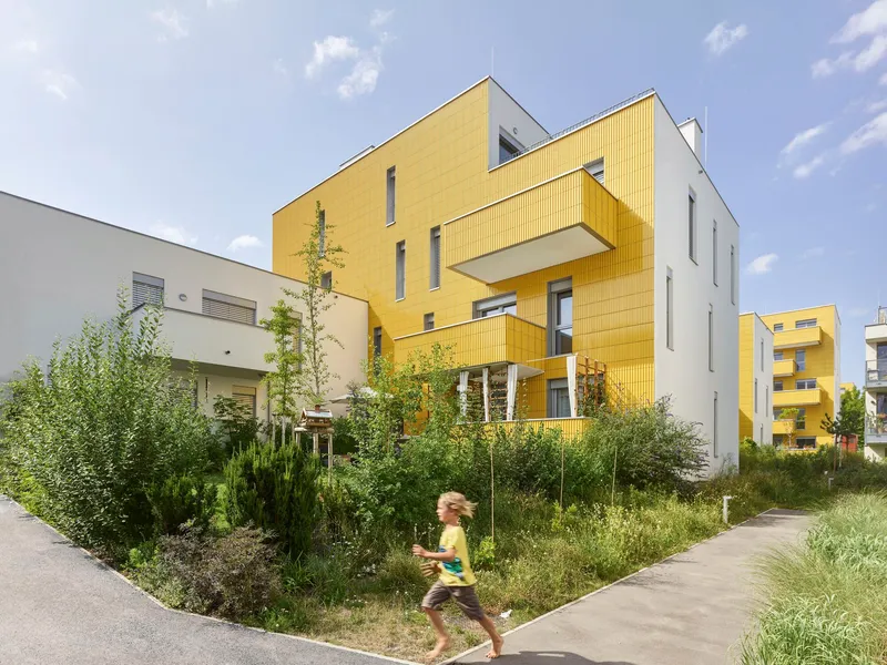 Yellow and white residential volumes with projecting balconies viewed across landscaped garden with running child