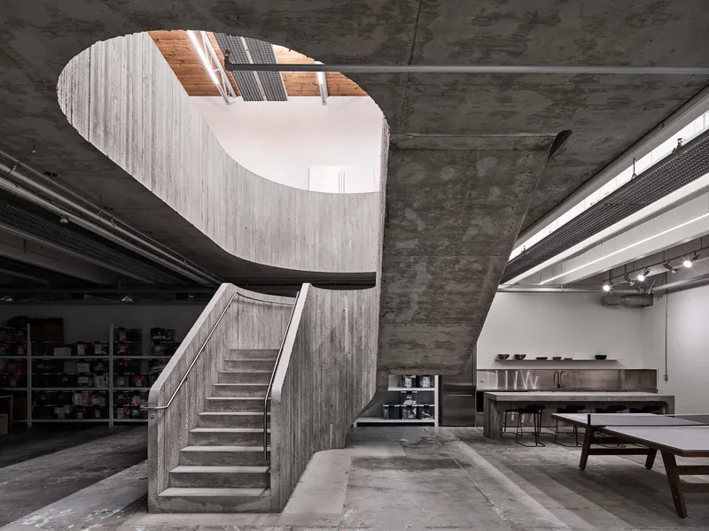 Curved concrete staircase with board-formed walls rising through an oval ceiling opening to natural light