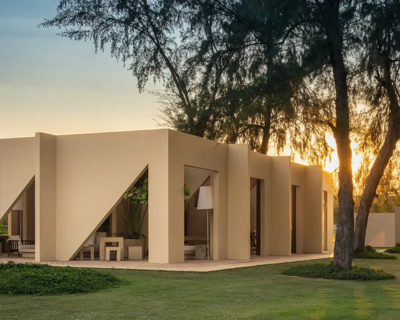 Folded facade with triangular openings framing furniture and plants at sunset beneath overhanging trees