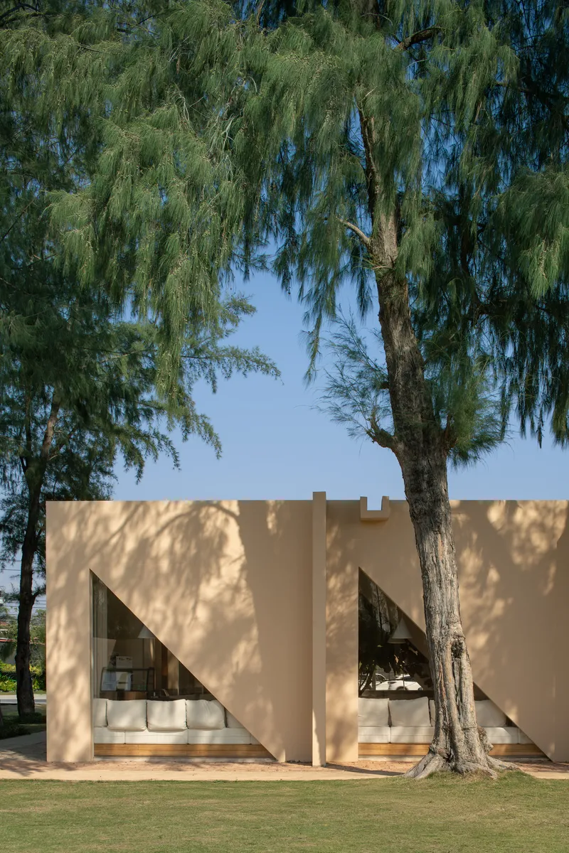 Cream stucco facade with triangular window openings beneath the dappled shade of pine trees