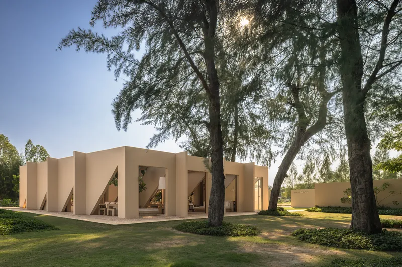 Folded facade with angled openings framing a covered porch beneath tall trees in afternoon light