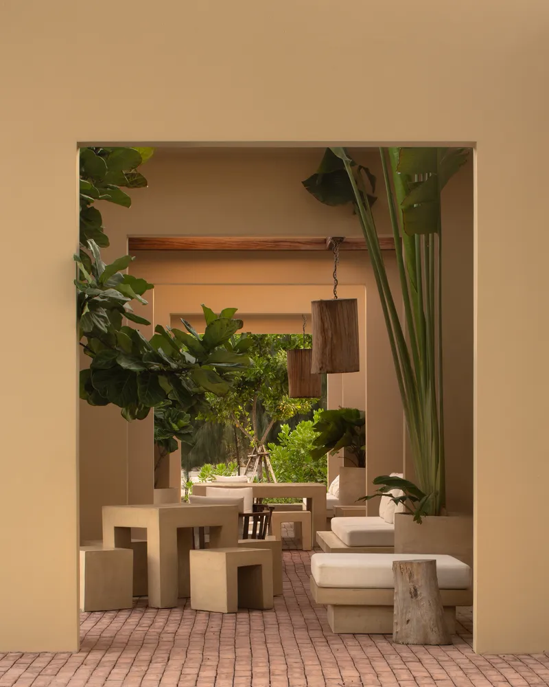Framed view through an opening showing dining furniture, hanging timber lights, and potted plants on brick pavers