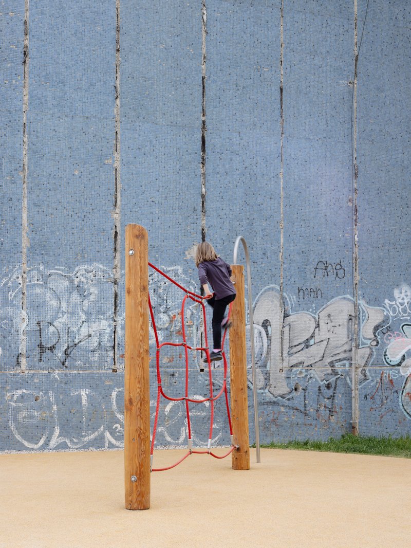 Child climbing red rope net structure on timber posts against a weathered blue tile wall