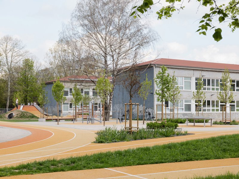 Schoolyard with orange rubber surfacing and planted beds between two gabled buildings under overcast skies