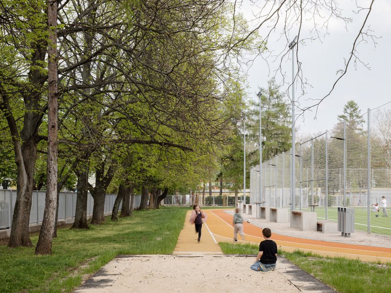Tree-lined path beside the sports court with figures running in dappled shade