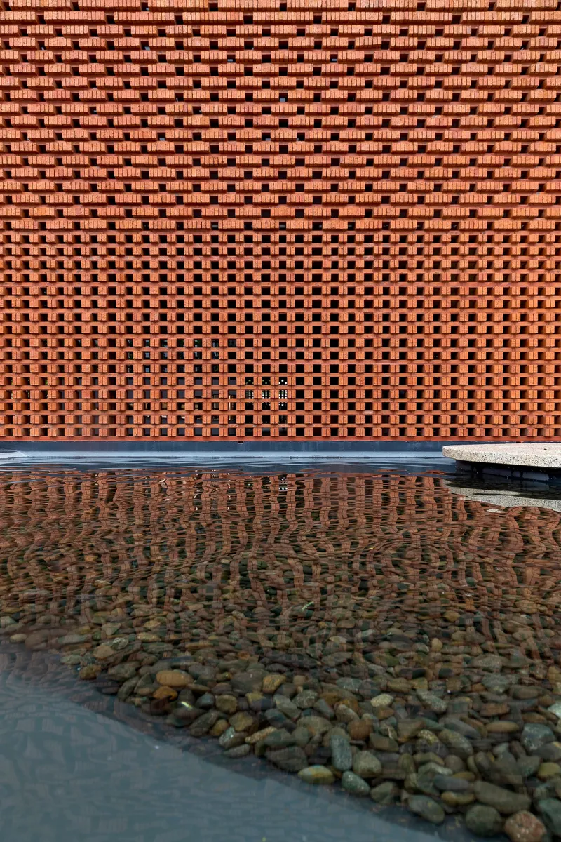 Perforated brick screen wall with varying aperture patterns reflected in shallow water pool with river stones