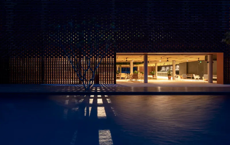 Perforated brick facade with a young tree casting shadows on the pool deck at dusk