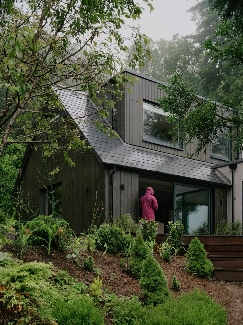 Figure in pink robe on planted slope below the dark timber and slate facade