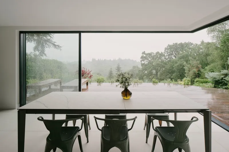 Dining table with marble top and dark chairs framing floor-to-ceiling glazing overlooking a misty landscape