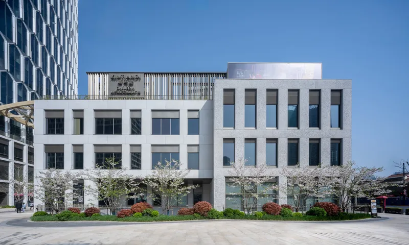 White precast facade with punched openings fronted by flowering trees and planted beds in the plaza