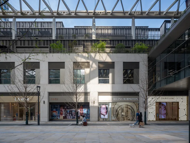White concrete facade with horizontal windows and street-level retail storefronts flanked by young trees in afternoon shadow