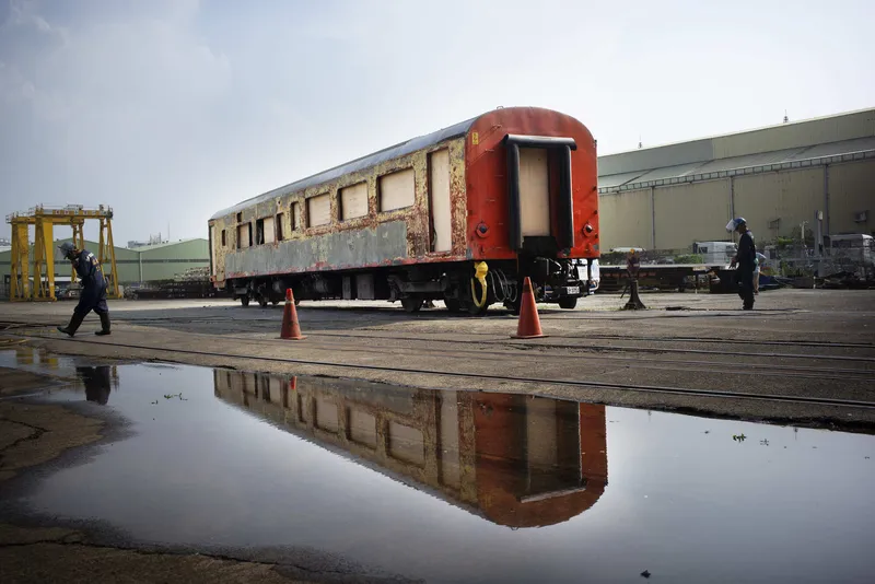 Weathered rail carriage on wet tarmac reflecting in a puddle with workers and traffic cones nearby
