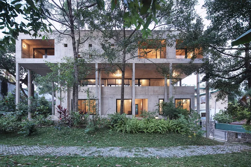 Three-storey concrete frame residence with glass openings surrounded by mature trees and planted lawn at dusk