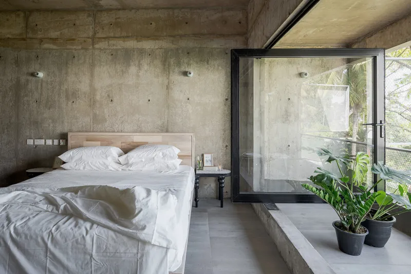 Bedroom with exposed concrete walls, timber headboard and black-framed glass door opening to planted balcony