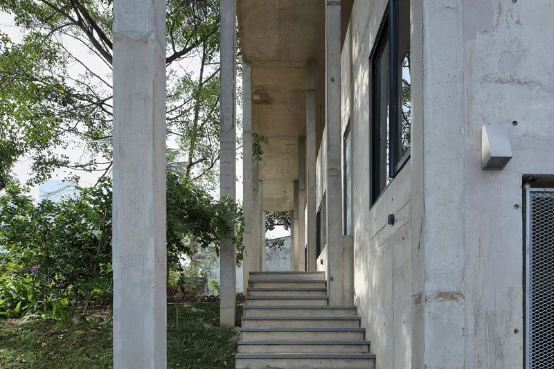 Exterior concrete stair rising between cylindrical columns toward a timber-clad soffit with trees beyond