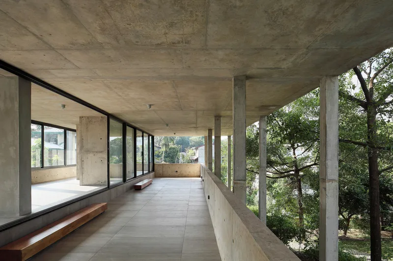 Long concrete corridor with exposed slab ceiling and timber bench along glazed wall facing trees