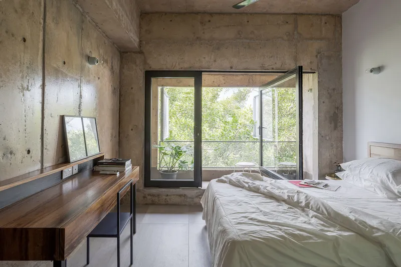 Bedroom with exposed concrete walls and ceiling, open casement window revealing greenery outside