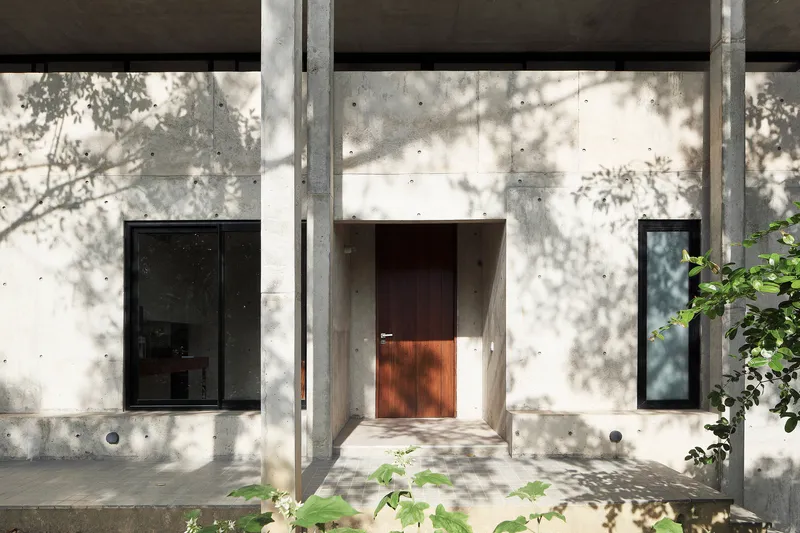 Concrete entrance facade with timber door and dappled tree shadows across the walls