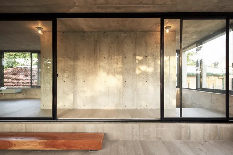 Interior view through glass partitions showing board-formed concrete walls with dappled sunlight from adjacent rooms