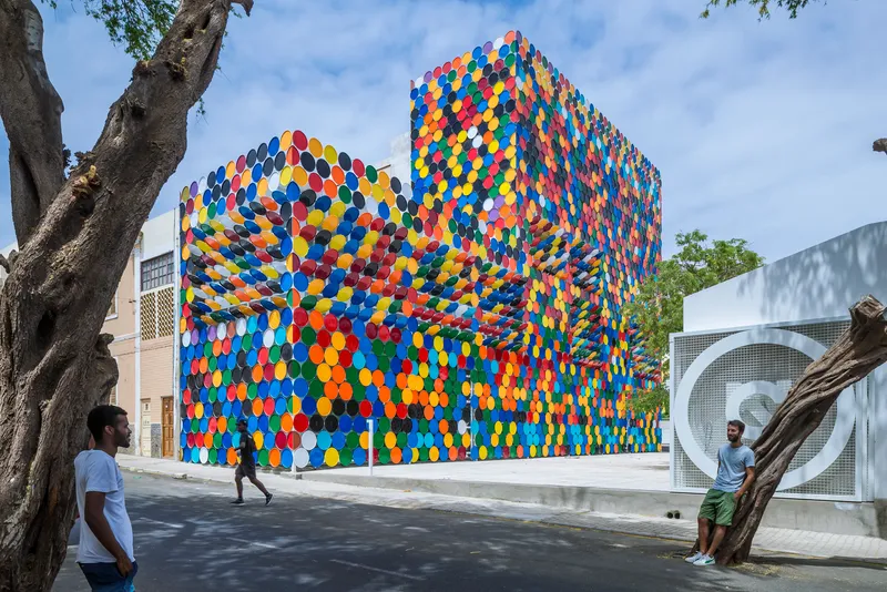 Street view of the facade clad in multicoloured circular discs beneath a tree and blue sky