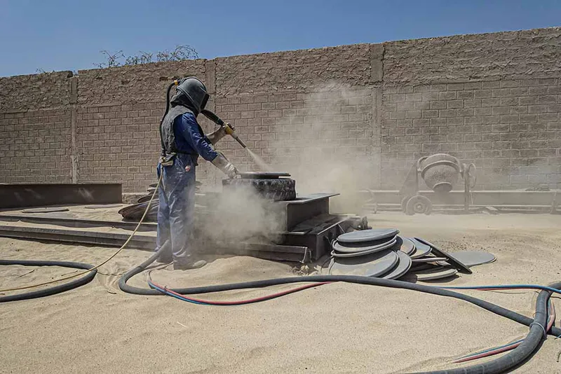 Worker sandblasting steel formwork in a dusty fabrication yard beside concrete block walls