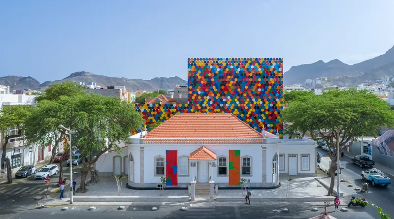 Aerial view showing the polychromatic disc facade rising behind a terracotta-roofed white building and green trees