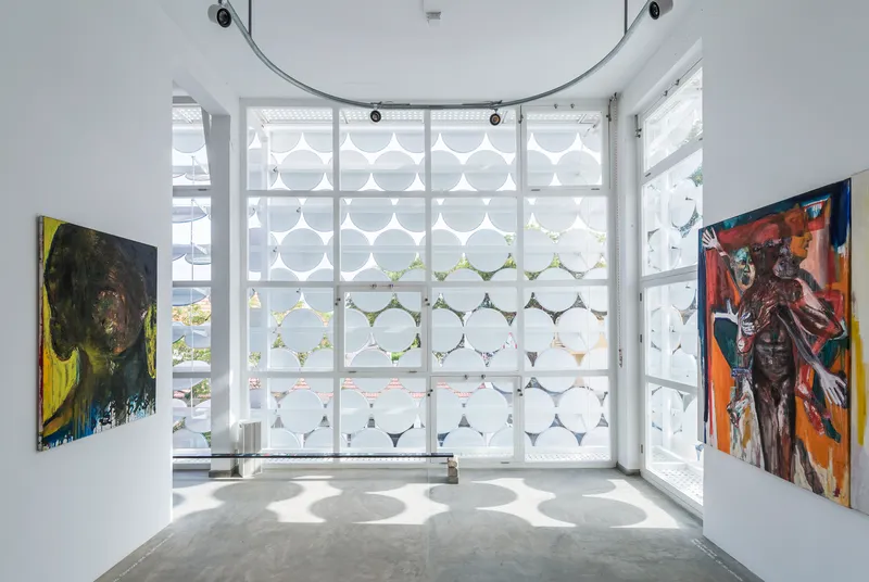 Gallery corner with floor-to-ceiling circular patterned screens filtering daylight between two paintings on white walls