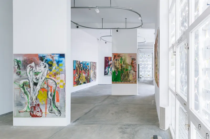Gallery space with curved track lighting and paintings displayed along white walls beside the translucent screen facade