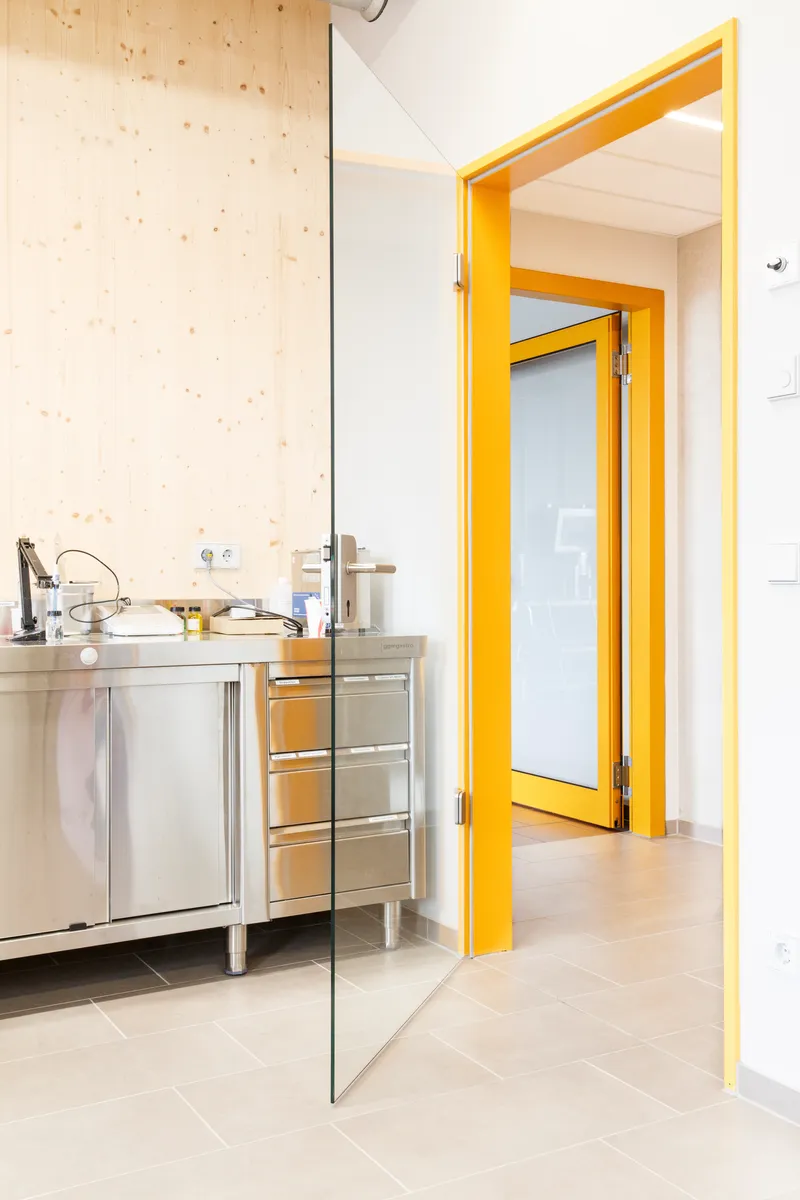 Yellow-framed doorway opening from tiled room with stainless steel counter into adjoining space with timber walls